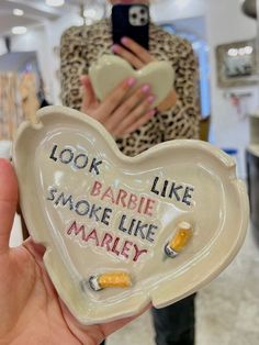 someone holding up a heart shaped ashtray with words written on it in front of a woman taking a selfie