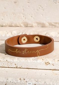 a brown leather bracelet with two gold studs on the clasp that says, love is strong