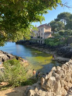 people are swimming in the water near some large rocks and trees, with a house on the other side