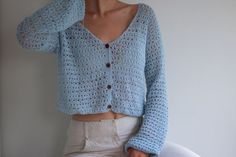 a woman wearing a blue crochet cardigan and white pants with her hands on her head