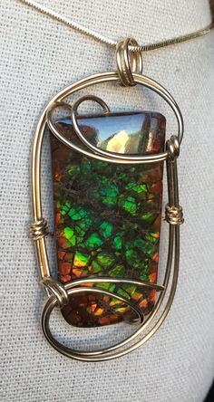 "This Ammolite pendant holds a freeform gemstone with no resin coating. The gem itself has a nice dragonskin pattern in green with teal overtones, and has a nice red edging. The wrap is 14K gold filled wire. The stone measures in at 4.0cm x 2.0cm with a total pendant size of 6.0cm x 2.8cm. Gold filled wire is more expensive than solid sterling silver wire, due to the high content of gold in it. By law, the bonded surface layer of gold must be one twentieth of the thickness of the wire to be considered gold filled. Therefore, it is next to impossible to wear off as plating often does. Gold filled is a very good alternative to solid gold, as it has the same wearability and visual appearance without the exorbitant cost. The pendant comes on an 18\" gold plated snake chain. Other lengths avail Resin Coating, Snake Chain, Silver Wire, Alex And Ani Charm Bracelet, Pendant Necklaces, Solid Gold, Gold Filled, Jewelry Necklace Pendant, Charm Bracelet