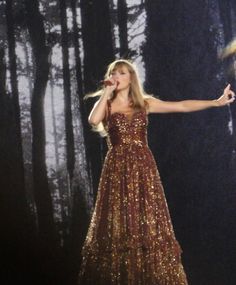 a woman in a long dress holding a microphone and performing on stage with trees behind her