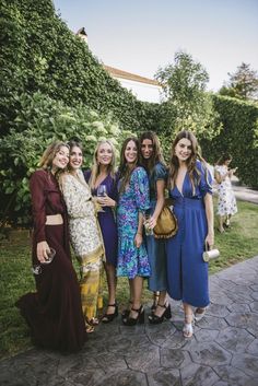 four women are posing for a photo in front of some bushes and trees, one is holding a purse