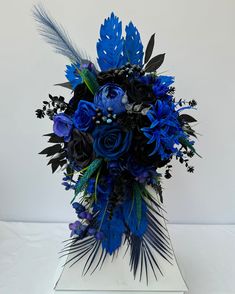 a bouquet of blue flowers and feathers in a square vase on a white tablecloth