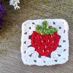 a crocheted square with a red strawberry on it and purple flowers in the background