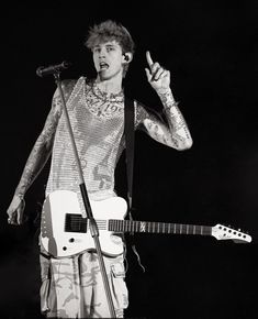 a man with tattoos on his arm holding a guitar and pointing to the side while standing in front of a microphone