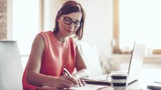 a woman sitting at a desk with a laptop and pen in her hand, writing