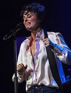 a woman singing into a microphone while wearing a white shirt and black tie with her hands in the air