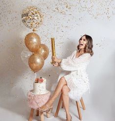 a woman sitting on a stool holding a cake and some balloons in front of her