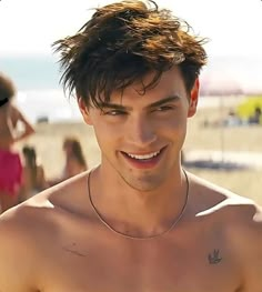 a shirtless man standing on the beach with people in the background and one person smiling at the camera