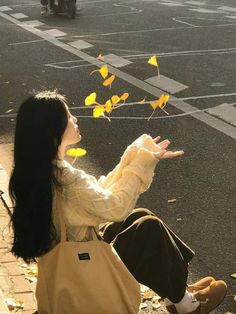 a woman is sitting on the curb with her hand out to catch some yellow leaves