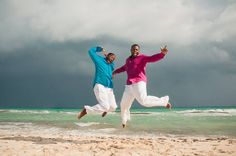 two people jumping in the air on a beach with storm clouds behind them and one person holding his arms out