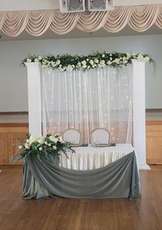 a table with white flowers and greenery on it in front of a curtained window