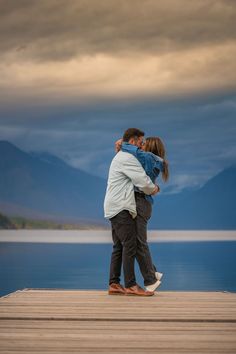 a man and woman embracing on a dock with mountains in the backgrouund