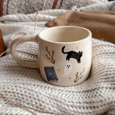 a coffee cup with a black cat on it sitting on a white blanket next to a brown bag