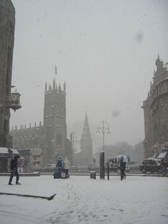 people are walking through the snow in front of some old buildings and clock tower on a snowy day