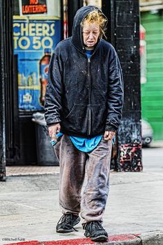 a man standing on the sidewalk in front of a building wearing a black jacket and blue pants