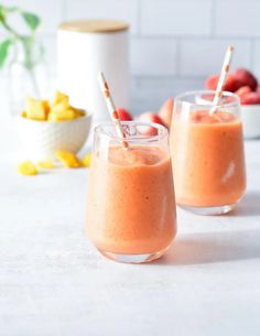 two glasses filled with orange smoothie on top of a white table next to bowls of fruit