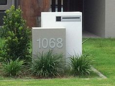 two mail boxes sitting in the grass next to some bushes and trees with a building behind them