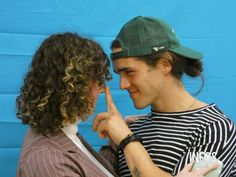 two people standing next to each other in front of a blue wall with one pointing at the other