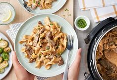 two hands holding plates with pasta and vegetables next to an oval casserole dish