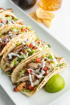 several tacos on a white plate with limes and guacamole in the background