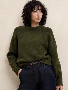 a woman with curly hair wearing a green sweater and jeans, leaning against a wall