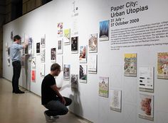 two men standing next to a white wall covered in pictures and papers, with one man leaning on the wall