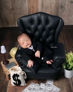 a baby in a black suit and tie is sitting on a chair next to money