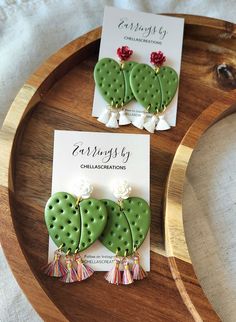 two green cactus earrings with tassels on top of a wooden tray next to a card