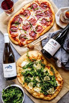 two pizzas with different toppings next to bottles of wine and glasses on the table