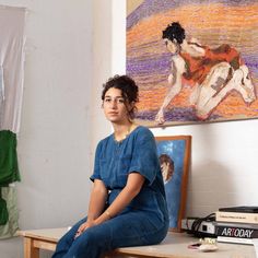 a woman sitting on top of a wooden bench in front of some paintings and books