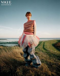 a woman standing on top of a grass covered field next to the ocean with her hands in her pockets