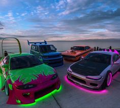 four different colored cars parked next to each other in front of the ocean at sunset