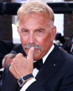 an older man in a tuxedo with a watch on his left hand and finger to his mouth