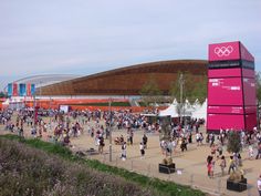 a large group of people walking around in front of an olympic sign and some buildings