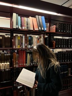 a woman standing in front of a bookshelf holding an open book and looking at it