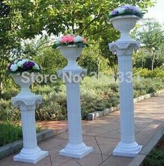 three white vases with flowers on them sitting in the middle of a sidewalk next to trees