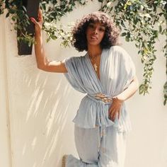 a woman standing in front of a white wall wearing a blue jumpsuit and holding her hand up