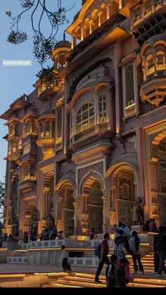 people walking in front of an ornate building at night with lights on the windows and balconies