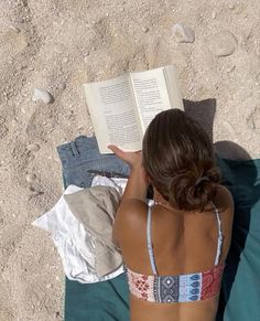 a woman reading a book while laying on top of a towel in the sand at the beach