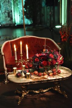 a table with candles and flowers on it in front of a red velvet couch at night