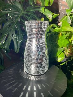 a silver vase sitting on top of a black table next to green plants and potted plants