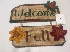 two welcome signs hanging from hooks on a white tablecloth covered surface with the words fall written in green, yellow and red