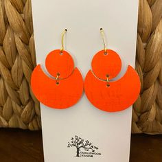 an orange pair of earrings is displayed in front of a white card and woven basket