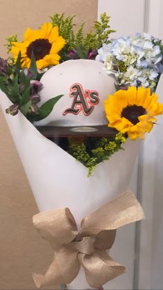 a baseball hat with flowers in it on top of a white vase filled with sunflowers and greenery