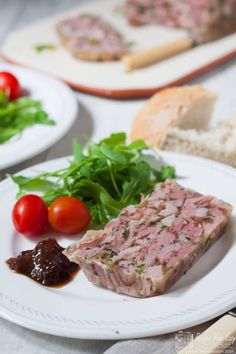 two plates with meat and vegetables on them next to bread, tomatoes and lettuce