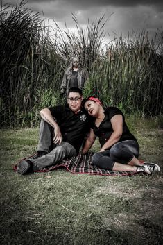 two people sitting on a blanket in front of some tall grass and one person standing behind them