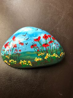 a painted rock sitting on top of a brown leather surface with flowers and birds in the sky