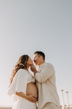 a pregnant couple kissing while standing on the beach